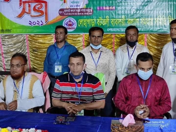 মঞ্চে অতিথিরা বসে আছেন, পেছনে কয়েকজন দাঁড়িয়ে; গলায় পরিচয়পত্রের ফিতা